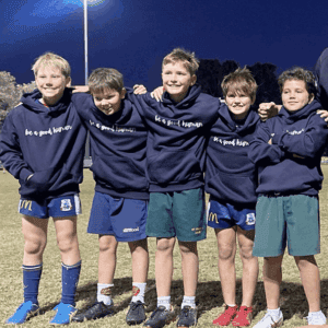 Five boys stand arm-in-arm on a grassy field at night, smiling in matching Kids Walk With Us Hoodies that say “be a good human.” Some wear shorts and sneakers, with lights glowing in the background.