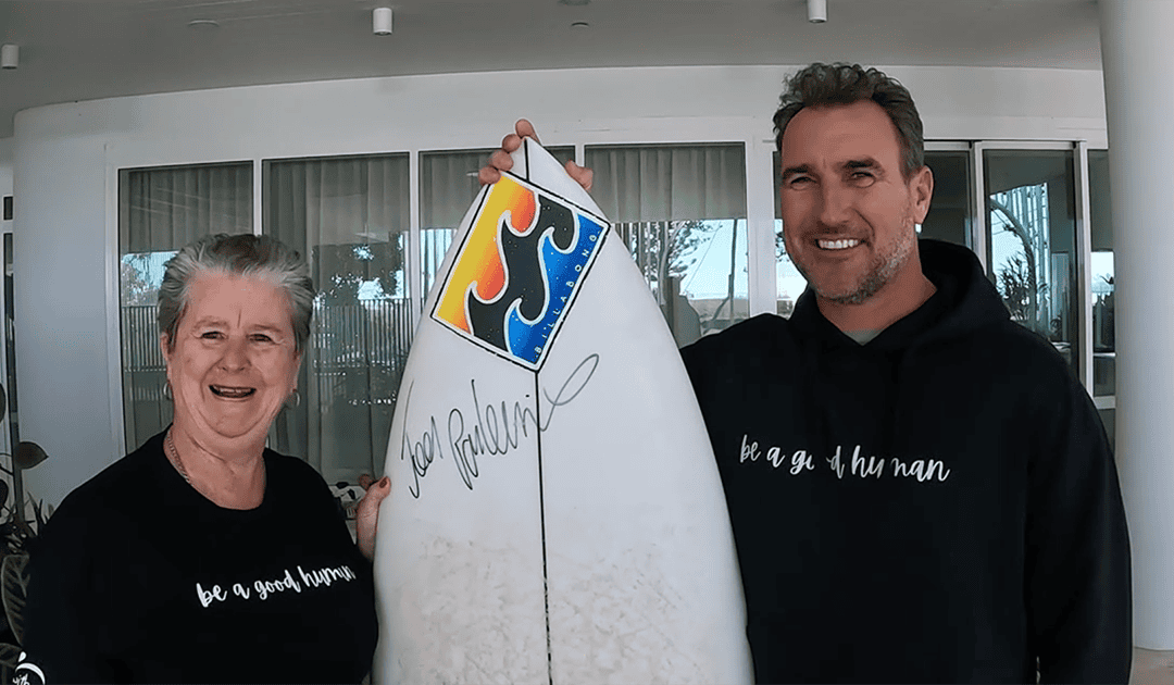 Two people smile while holding a surfboard with a colorful logo and signature. Both wear black shirts; the woman’s says “be a good human,” and the man's says “be a good hu man.” Indoors, with large windows in the background.
