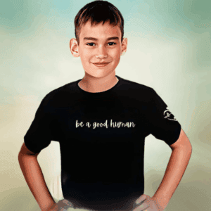 A smiling boy stands hands on hips, wearing the Kids Walk With Us Tee—a black T-shirt with “be a good human” on the front. The softly blurred background features light green and beige tones.