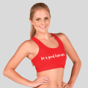 A smiling woman with long blonde hair in a ponytail poses with her hands on her hips, wearing the red "Ladies Walk With Us Stretch Crop Top" featuring "be a good human" in white script. The background is plain light gray.
